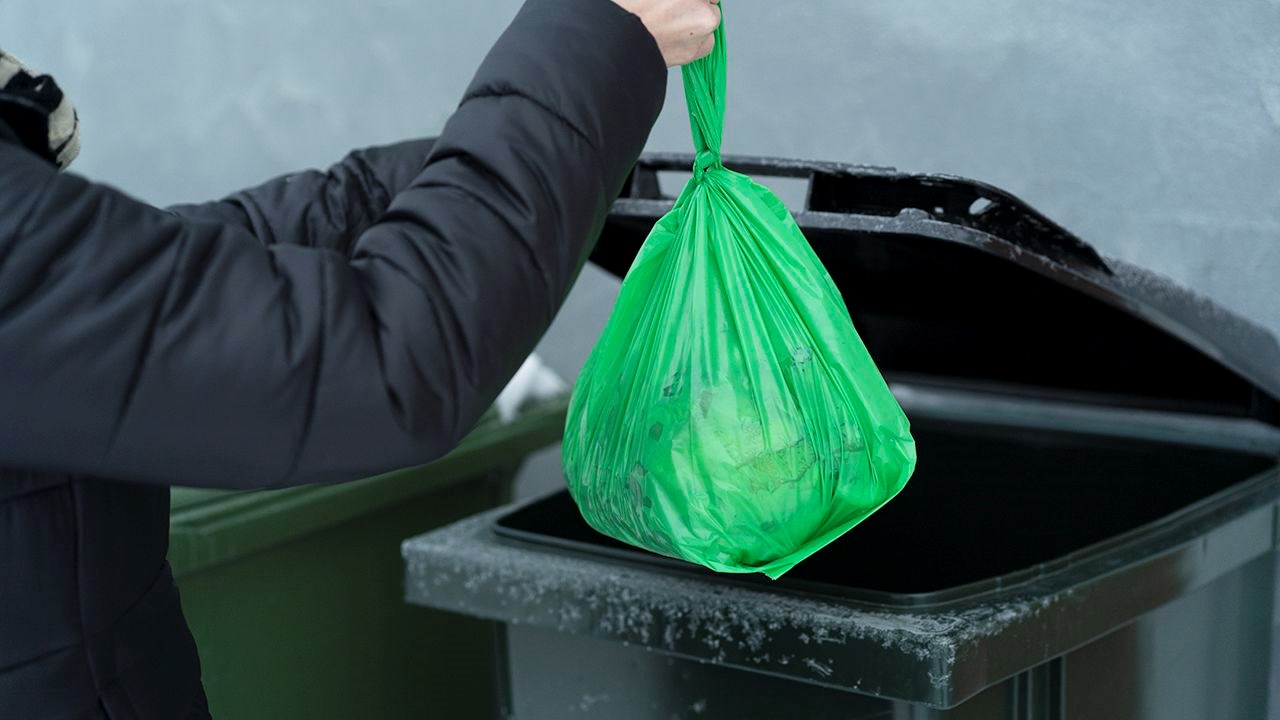 Bilden visar en person som slänger en grön påse i ett sopkärl.